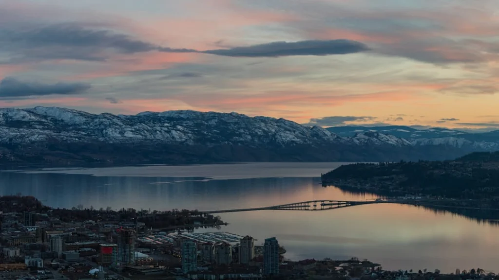 Panoramisch uitzicht op Kelowna met een brug over het meer, besneeuwde bergen en een kleurrijke avondlucht