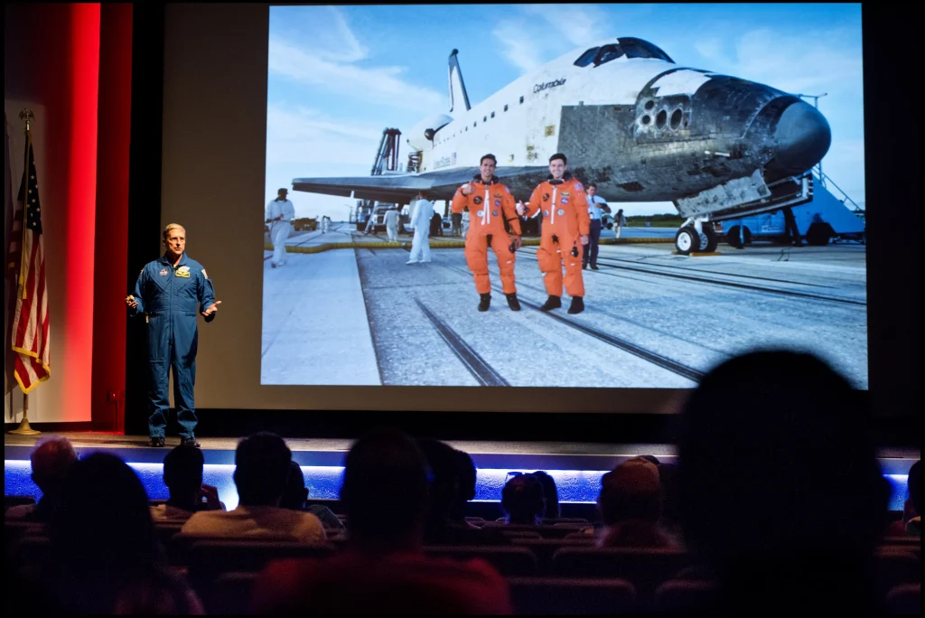 Astronaut in blauw pak vertelt op een podium over een ruimtevaartmissie naast een grote foto van een spaceshuttle