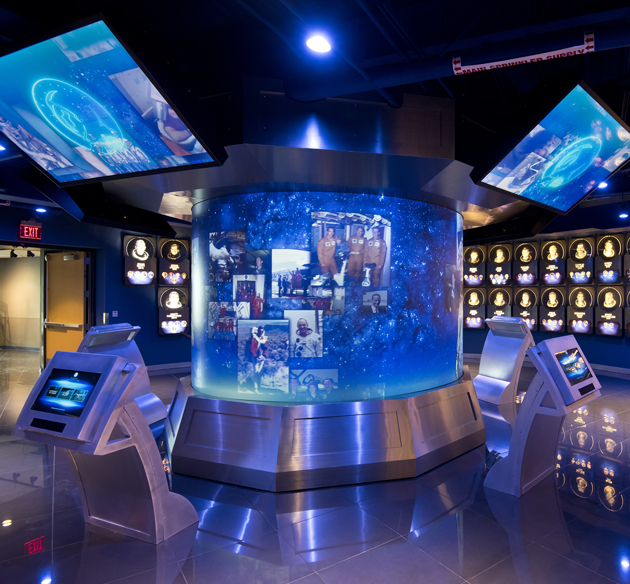 Blauw verlichte ronde vitrine met historische astronautenfoto's, omringd door interactieve schermen in een donkere museumzaal