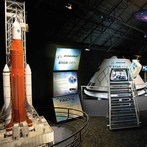 Model van een grote raket naast een Boeing Starliner capsule met informatiepanelen in een donkere tentoonstellingsruimte