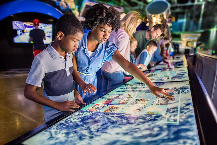 Twee kinderen wijzen naar een groot interactief tafelscherm met een wereldkaart terwijl andere bezoekers meekijken