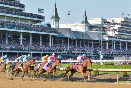 Jockeys op galopperende paarden razen door de bocht van de renbaan van Churchill Downs in Kentucky, met volle tribunes en de kenmerkende torentjes op de achtergrond