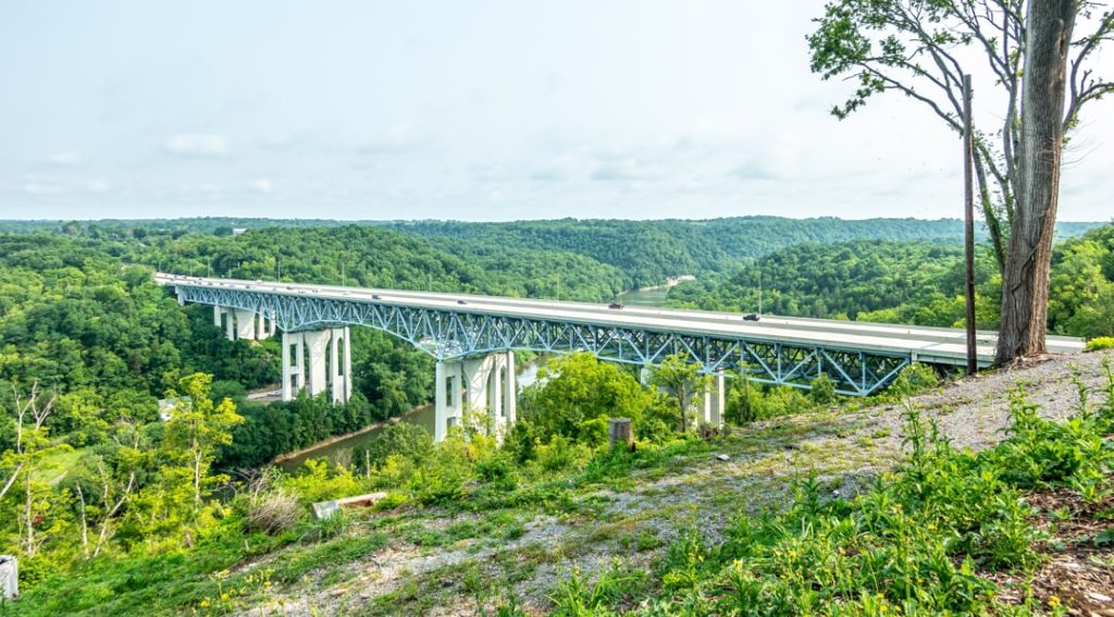 Lange stalen vakwerkbrug die over een diep beboste vallei loopt, gezien vanaf een heuvel met uitzicht op de rivier