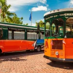 Twee oranje met groene hop-on hop-off trolleybussen staan geparkeerd op een geplaveide straat in Key West onder een zonnige hemel