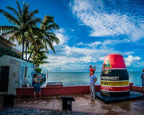 Toeristen fotograferen elkaar bij de rood zwart gestreepte Southernmost Point boei aan zee onder palmbomen in Key West