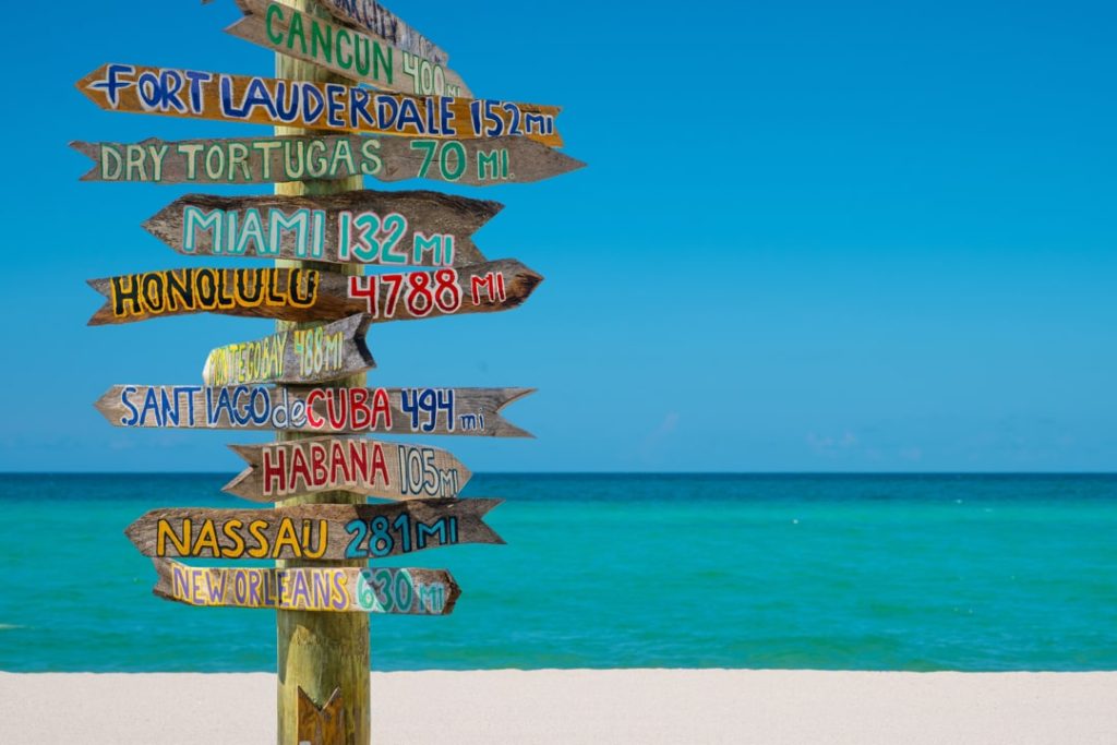 Houten paal met kleurrijke pijlen die de afstand aangeven naar steden als Havana, Miami en Nassau op een wit strand