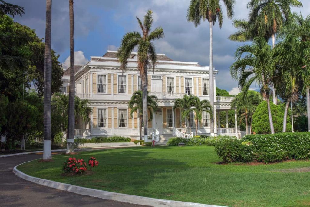 Witte koloniale villa Devon House in Kingston omgeven door hoge palmbomen, een verzorgd gazon en bloeiende rode struiken