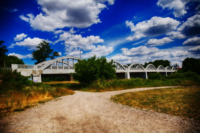 Wit zandpad dat leidt naar een lange stalen boogbrug over een rivier, met droog gras en struiken eromheen bij Kitchener