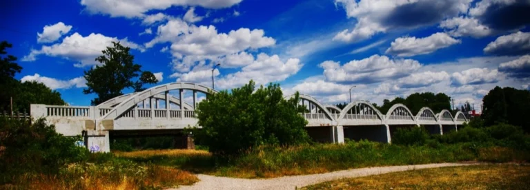Lange witte stalen boogbrug met meerdere bogen die over een rivier loopt, omgeven door struiken en gras bij Kitchener