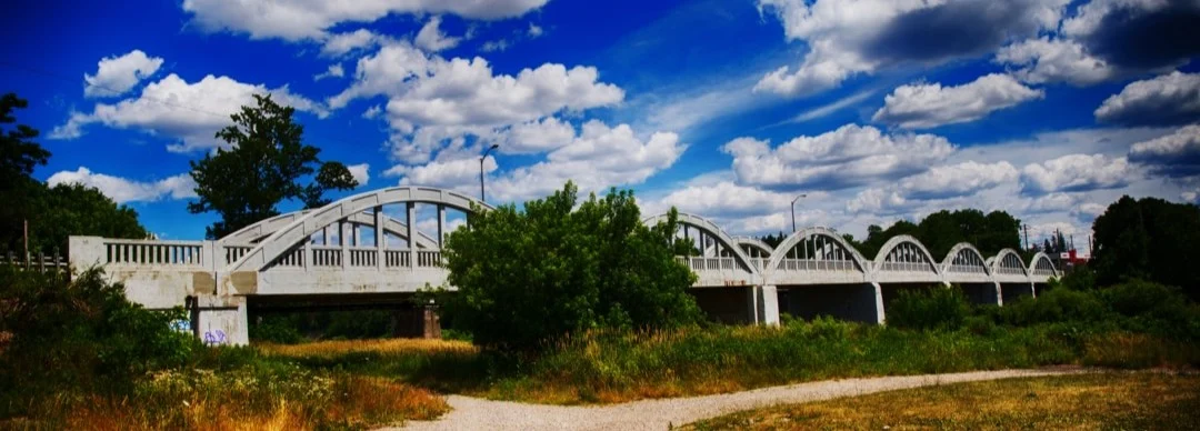 Lange witte stalen boogbrug met meerdere bogen die over een rivier loopt, omgeven door struiken en gras bij Kitchener