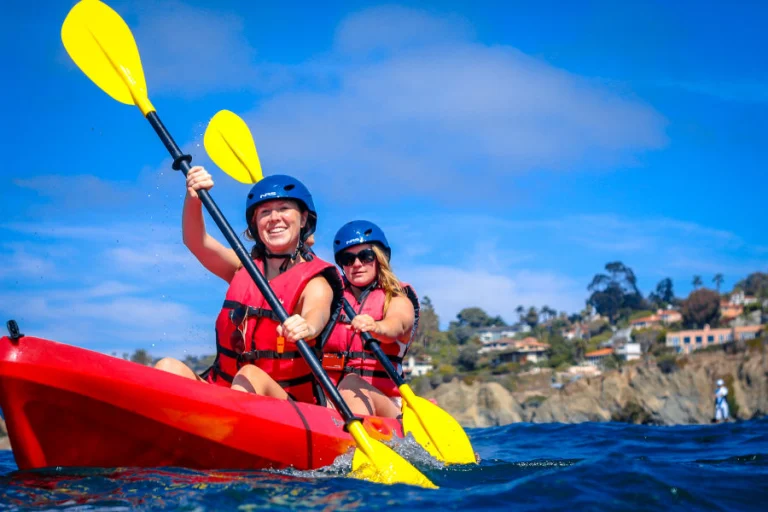 Twee vrouwen met blauwe helmen en reddingsvesten peddelen in een rode kajak op zee bij La Jolla, met de kust en huizen op de achtergrond