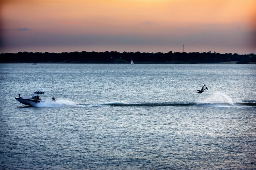 Wakeboarder springt hoog boven het water achter een speedboot op Lake Grapevine, met een oranje avondlucht op de achtergrond