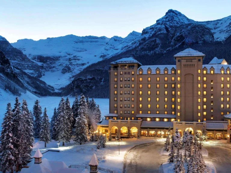 Een groot hotel met verlichte ramen staat in de schemering tussen besneeuwde sparren bij Lake Louise, omringd door hoge besneeuwde bergen