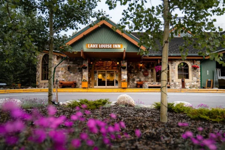 Buitenaanzicht van de stenen entree van Lake Louise Inn met een groen dak, omringd door bomen en paarse bloemen