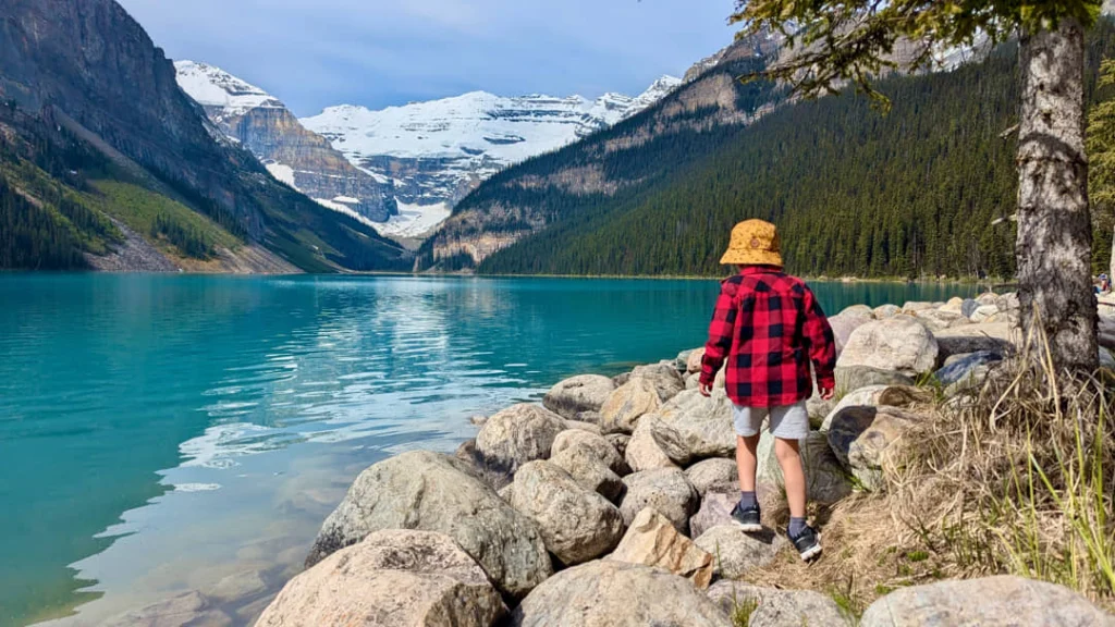 Jongen in rode ruitjesjas op rotsen aan de oever van Lake Louise met besneeuwde bergen op de achtergrond