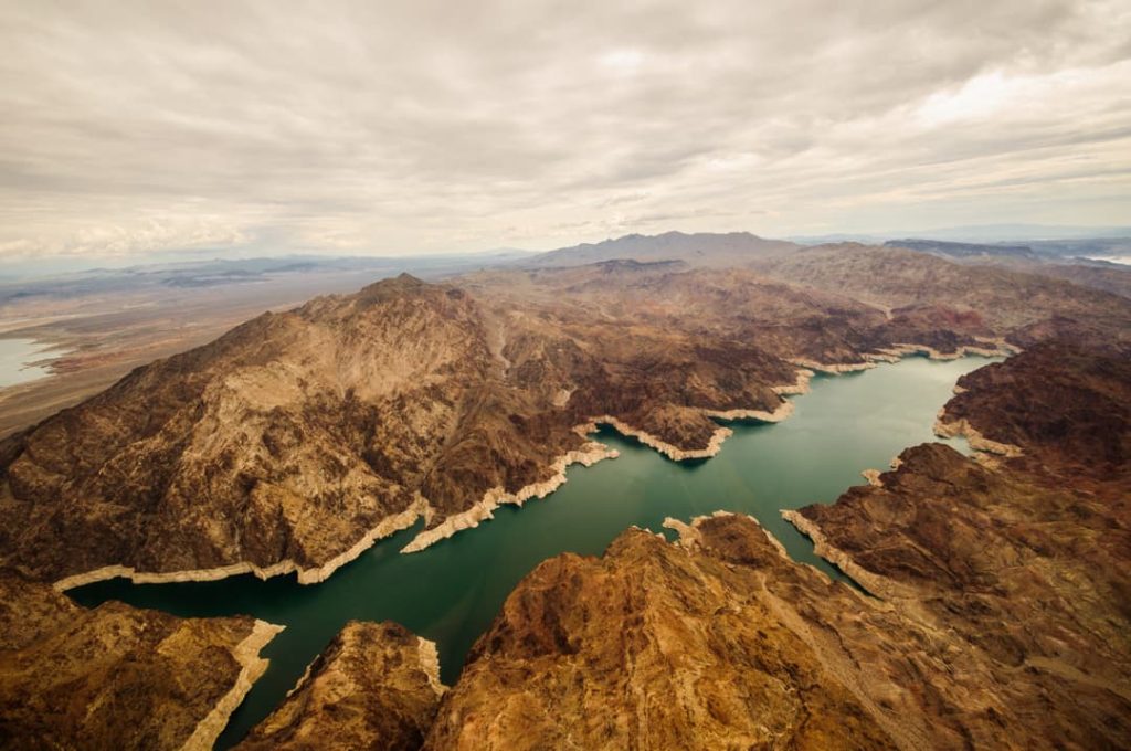 Vogelvluchtperspectief op het groenblauwe water van Lake Mead dat zich slingert tussen bruine, rotsachtige bergen onder een bewolkte lucht