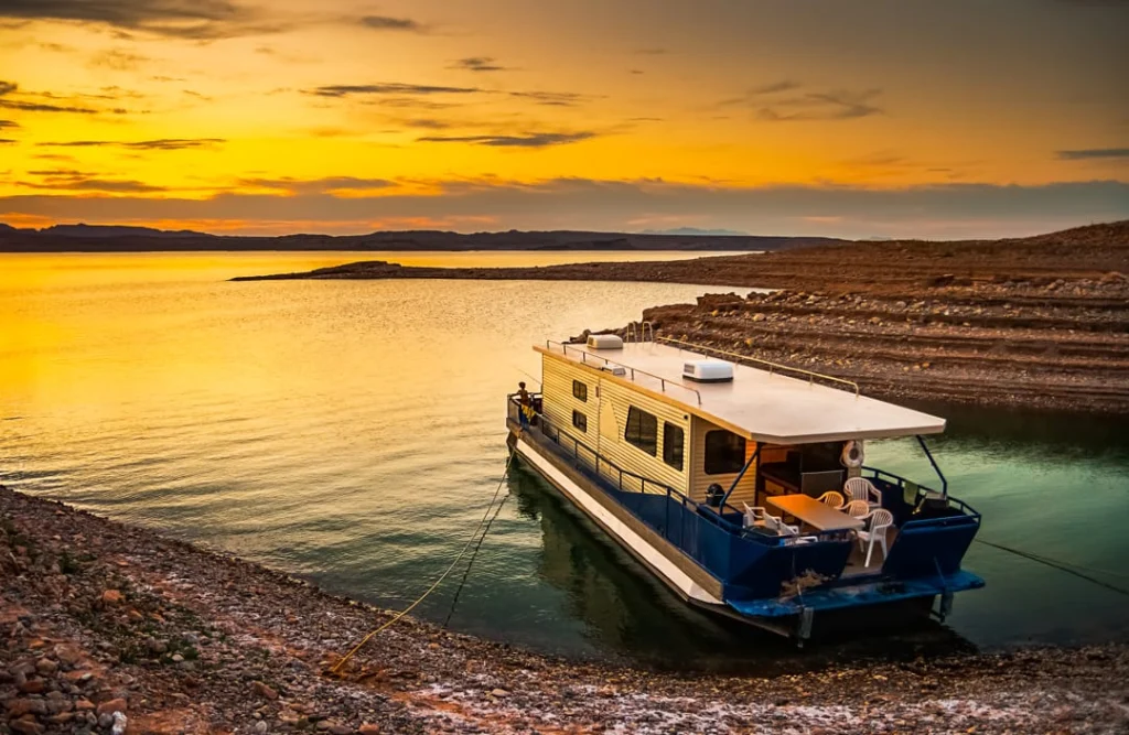 Grote woonboot ligt afgemeerd aan een rotsachtige oever van Lake Mead terwijl de lucht oranje kleurt bij zonsondergang