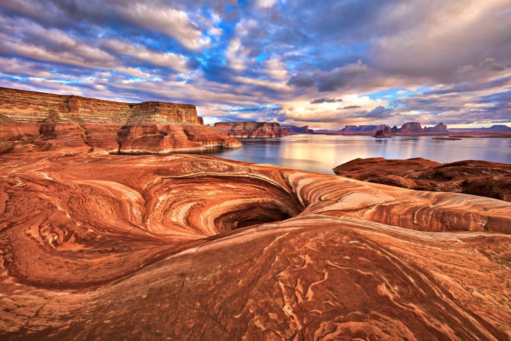 Golvende, gestreepte rode zandsteenrotsen op de voorgrond met het rustige water van Lake Powell en rotseilanden in de verte