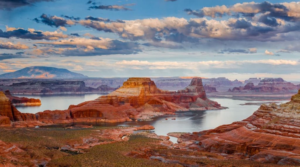 Wijds uitzicht over de rode rotseilanden en inhammen van Lake Powell met een verre vulkanische berg onder een bewolkte lucht