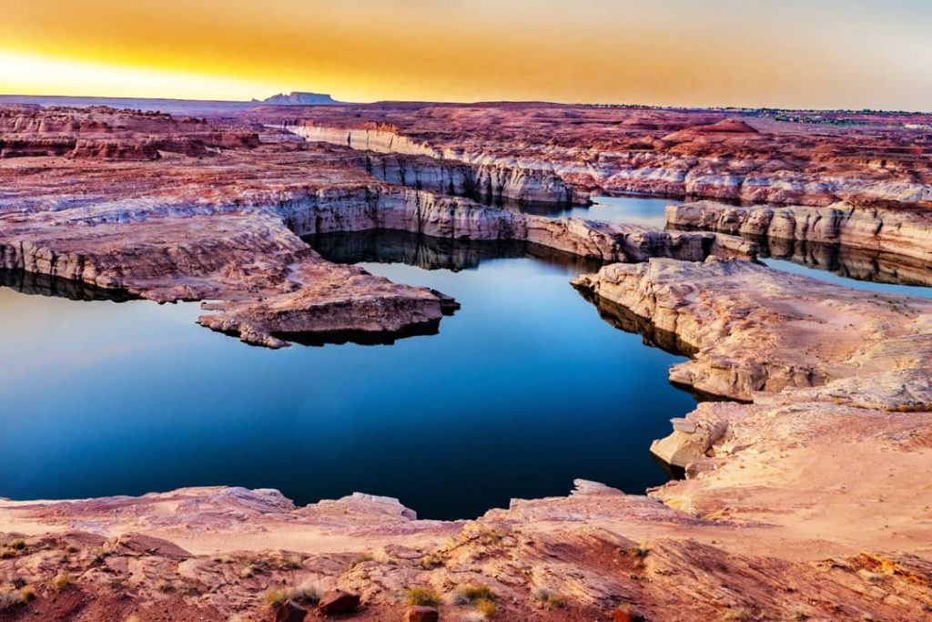 Rotsachtige canyonwanden van Lake Powell weerspiegelen in het rustige blauwe water onder een oranje avondlucht