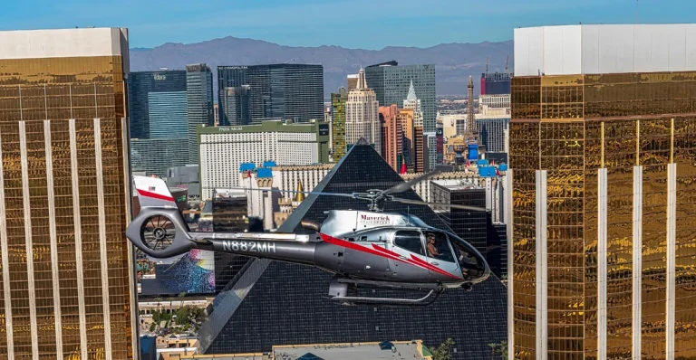 Een zilverkleurige helikopter van Maverick vliegt tussen hoge glazen wolkenkrabbers met de Las Vegas Strip en bergen op de achtergrond