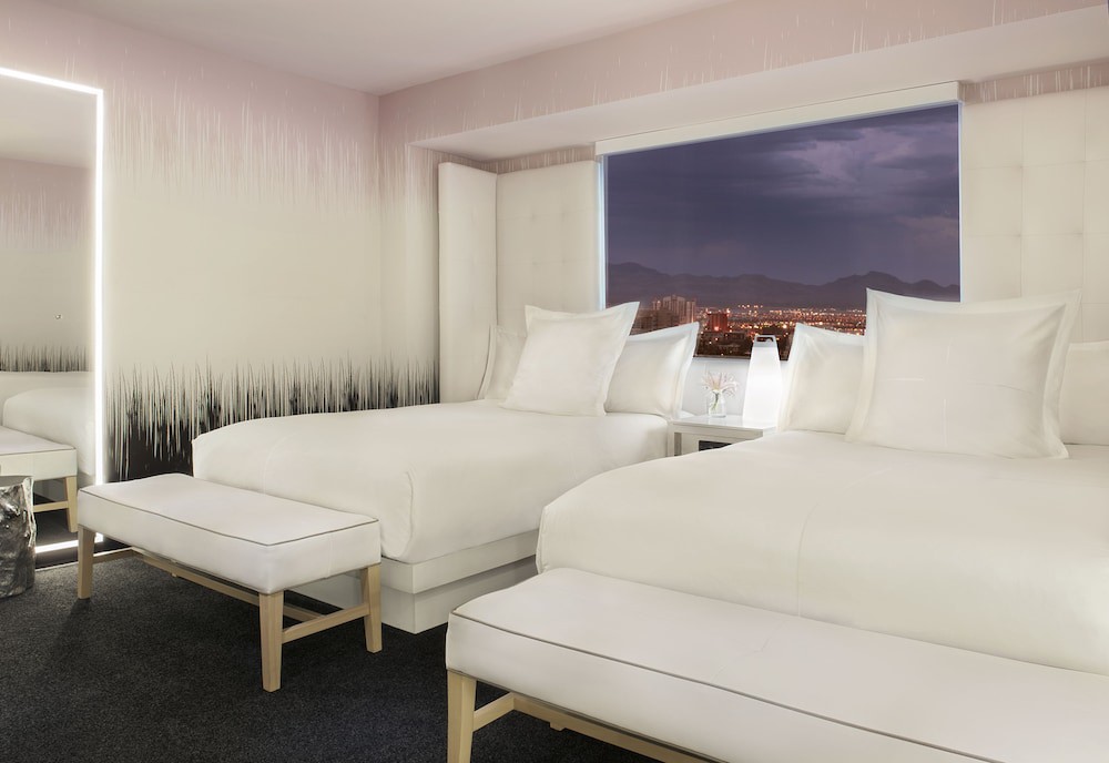 Twee witte bedden in een hotelkamer met uitzicht door het raam op de verlichte skyline van Las Vegas in de avond