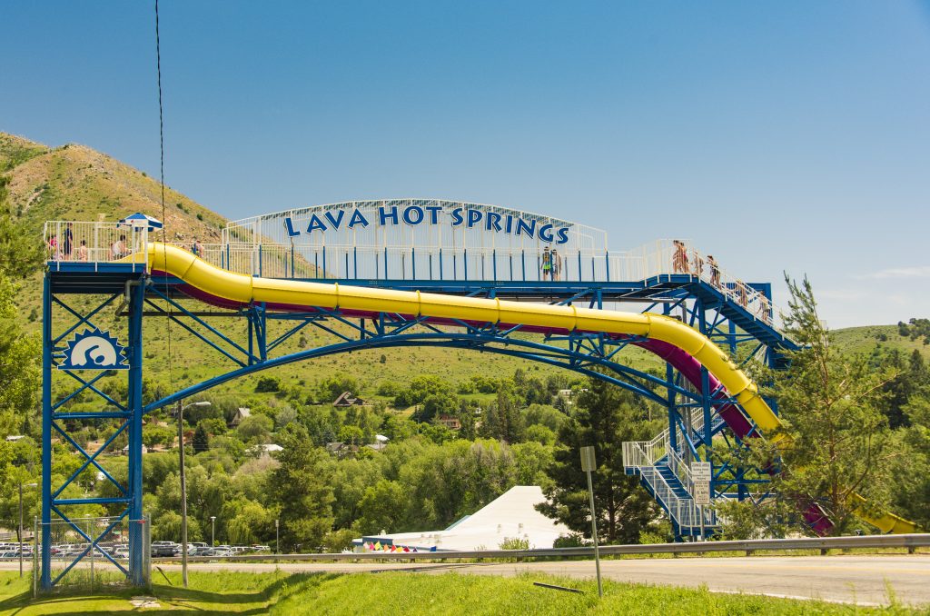 Lange gele waterglijbaan op een blauw frame met het opschrift Lava Hot Springs, tegen een groene heuvel