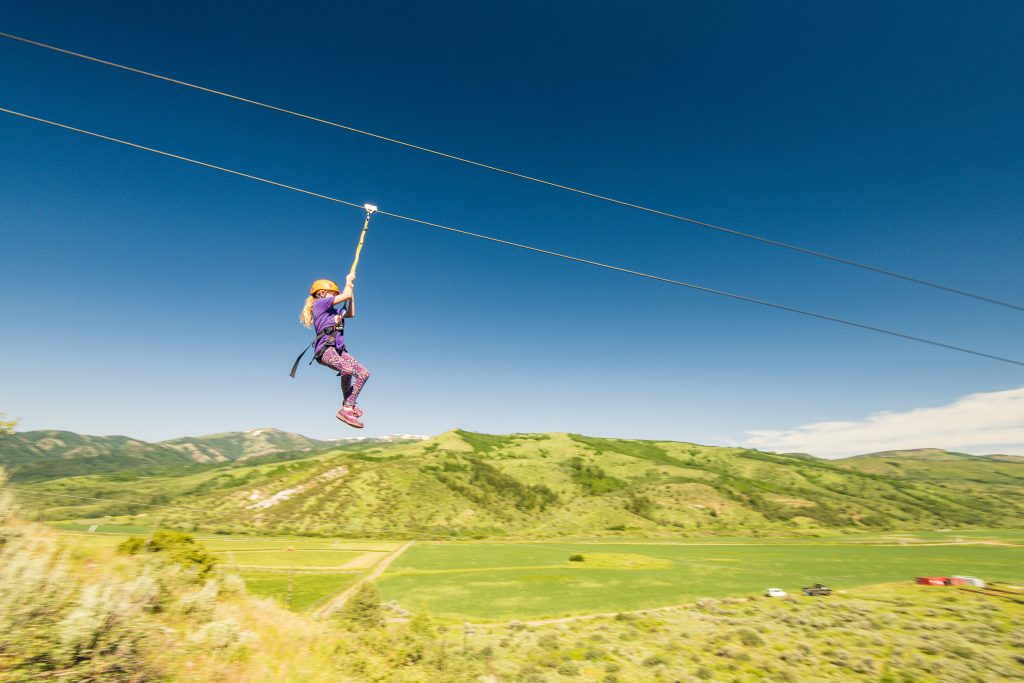 Persoon in een tuigje glijdt aan een zipline hoog boven een groen dal met glooiende heuvels op de achtergrond