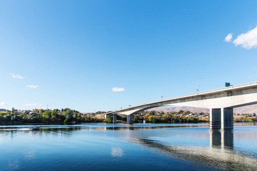 Lange betonnen brug overspant een brede rivier onder een strakblauwe lucht, met heuvels en bebouwing langs de oever