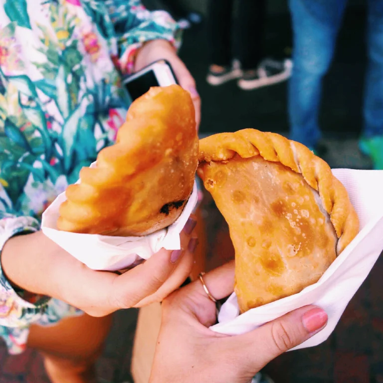 Twee handen houden elk een krokant gebakken empanada vast in een wit servet tijdens een culinaire wandeling door Little Havana