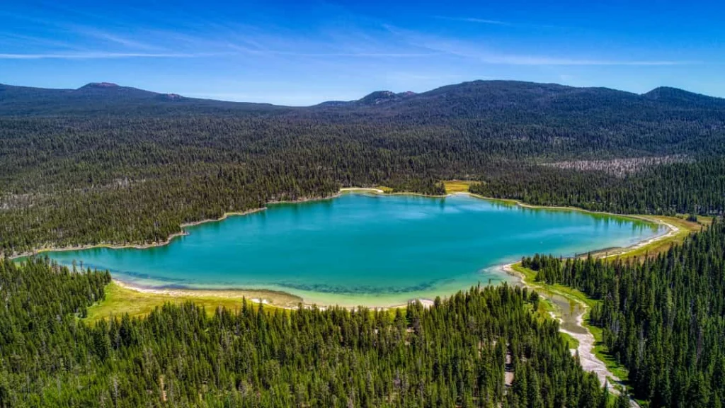 Luchtfoto van een turquoise gekleurd meer in Oregon volledig omgeven door dicht naaldbos onder een blauwe hemel