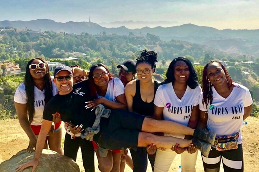 Groep vrouwen in gelijke t-shirts en een man tillen samen een vrouw op tijdens een wandeling met uitzicht over de heuvels van Los Angeles