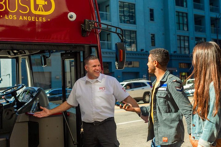 Glimlachende chauffeur van een rode sightseeingbus begroet twee toeristen naast de bus op straat in Los Angeles