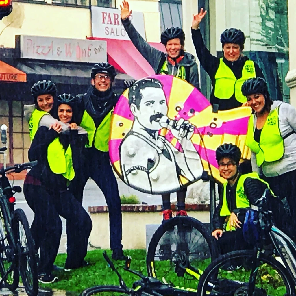 Groep fietsers met helmen en fluorescerende hesjes poseert lachend bij een kleurrijke muurschildering van een zingende man met microfoon