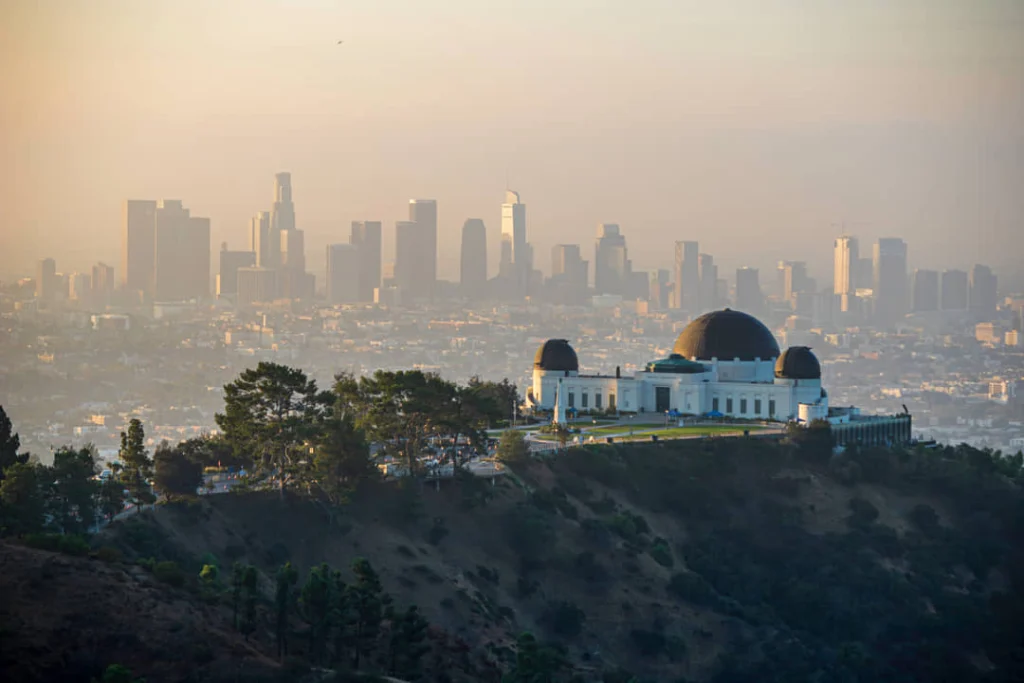 De witte koepels van Griffith Observatory op een heuvel met bomen, met de wazige skyline van Los Angeles op de achtergrond