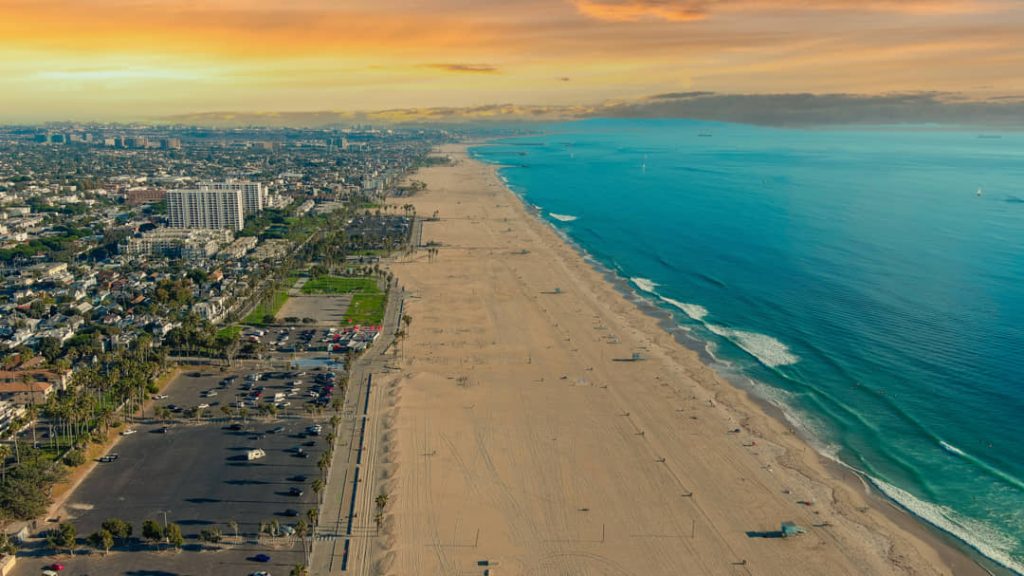 Luchtfoto van een breed zandstrand langs de Pacifische kust met turquoise water en hoogbouw van een kustplaats in de verte bij Los Angeles