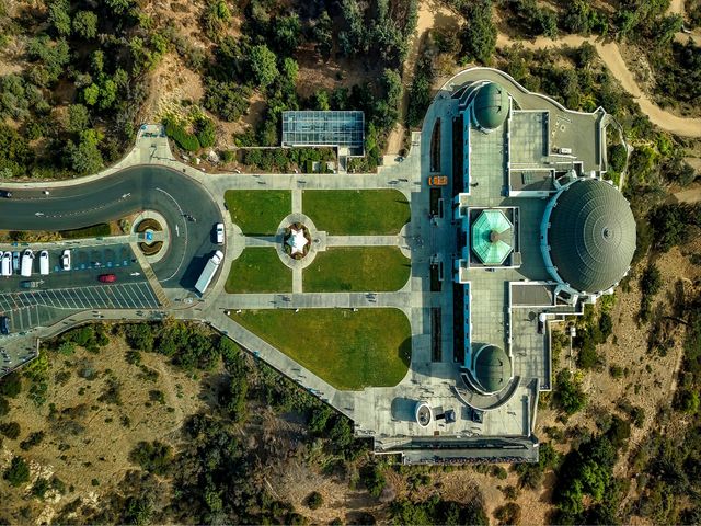 Luchtfoto recht van boven op het gebouw en de tuinen van het Griffith Observatory met een rondweg en parkeerplaats ernaast