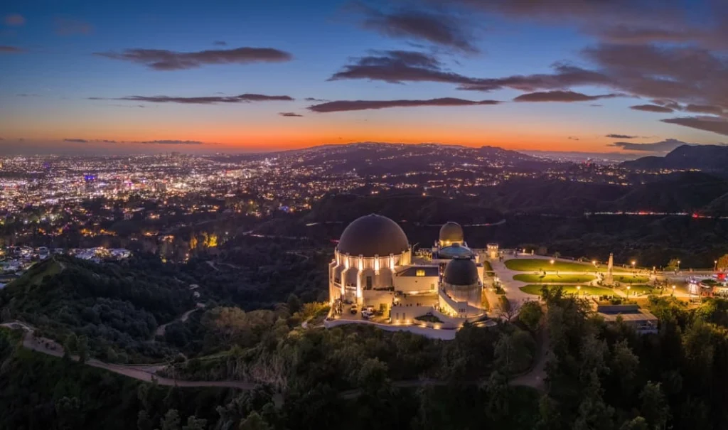 Luchtfoto van de koepels van Griffith Observatory op een heuvel met de verlichte skyline van Los Angeles op de achtergrond bij schemering