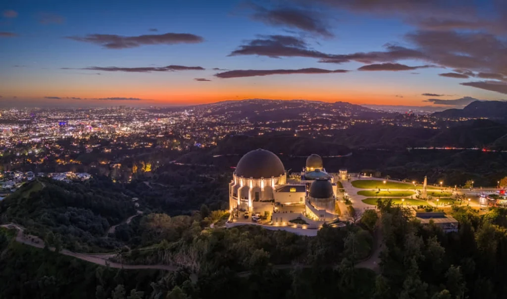 Luchtfoto van de verlichte koepels van Griffith Observatory op een heuvel met de skyline van Los Angeles op de achtergrond bij schemering