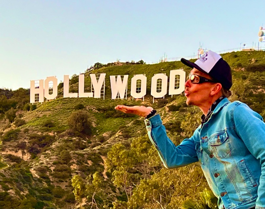 Een man in spijkerjasje en pet poseert met een kushandje voor het Hollywood-bord op de heuvel in Los Angeles
