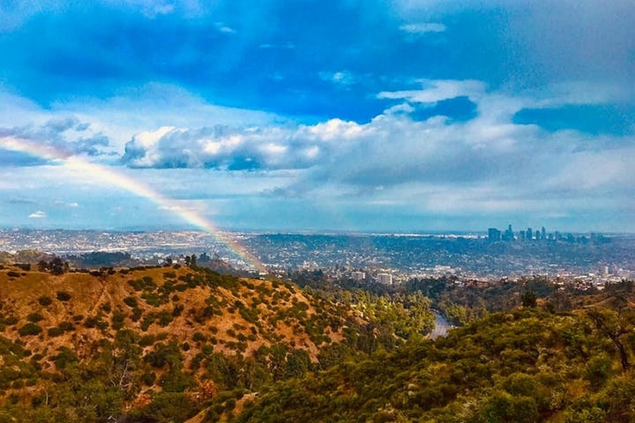 Een regenboog boven groene heuvels met in de verte de skyline van Los Angeles onder een bewolkte lucht