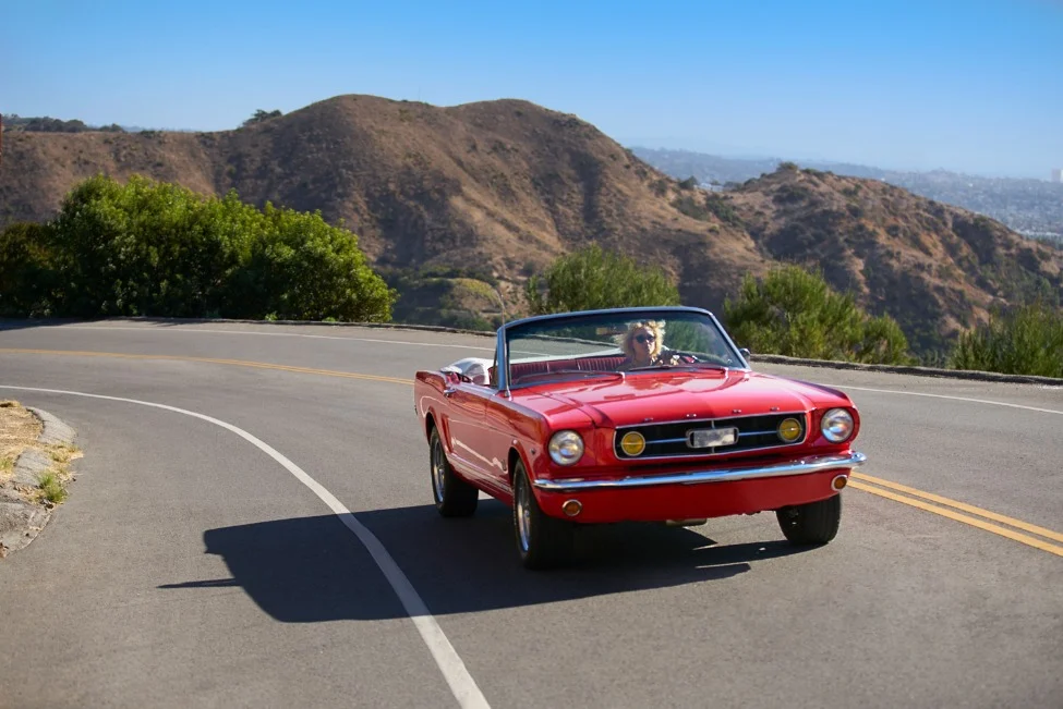 Een rode klassieke Ford Mustang cabriolet rijdt over een bochtige weg in de heuvels bij Los Angeles met de stad op de achtergrond