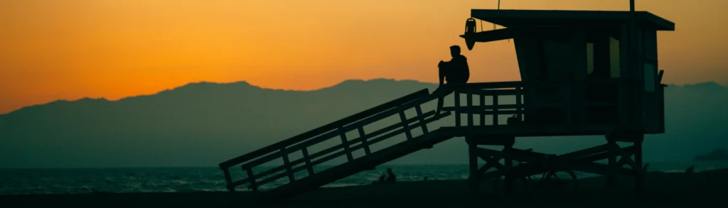 Silhouet van een persoon die op de reling van een strandwachtershutje zit terwijl de zon oranje ondergaat boven Venice Beach in Los Angeles