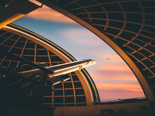 Binnenaanzicht van een open observatoriumkoepel met een telescoop tegen een roze en oranje avondlucht