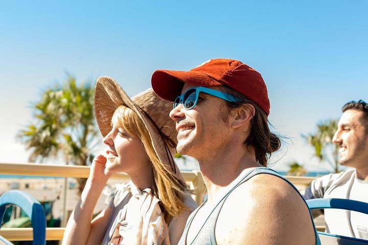Man en vrouw met zonnebril en hoed zitten op de bovenverdieping van een open sightseeingbus tijdens een tour in Los Angeles