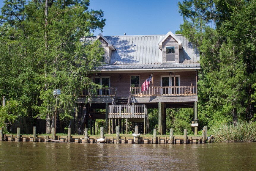 Grijs houten huis met veranda en Amerikaanse vlag gebouwd op palen boven het water van een bayou, omringd door bomen