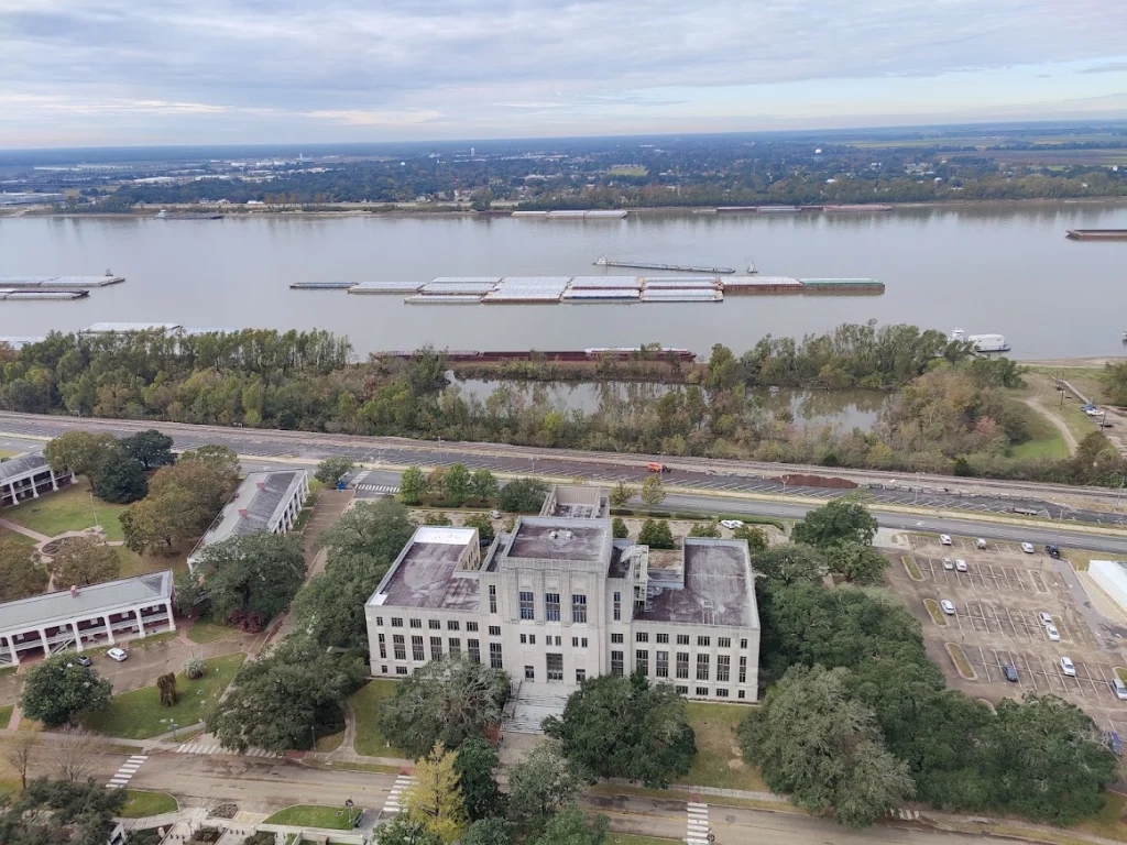 Luchtopname van een verweerd wit gebouw met vlak dak naast een snelweg met daarachter de brede Mississippi rivier met sleepboten
