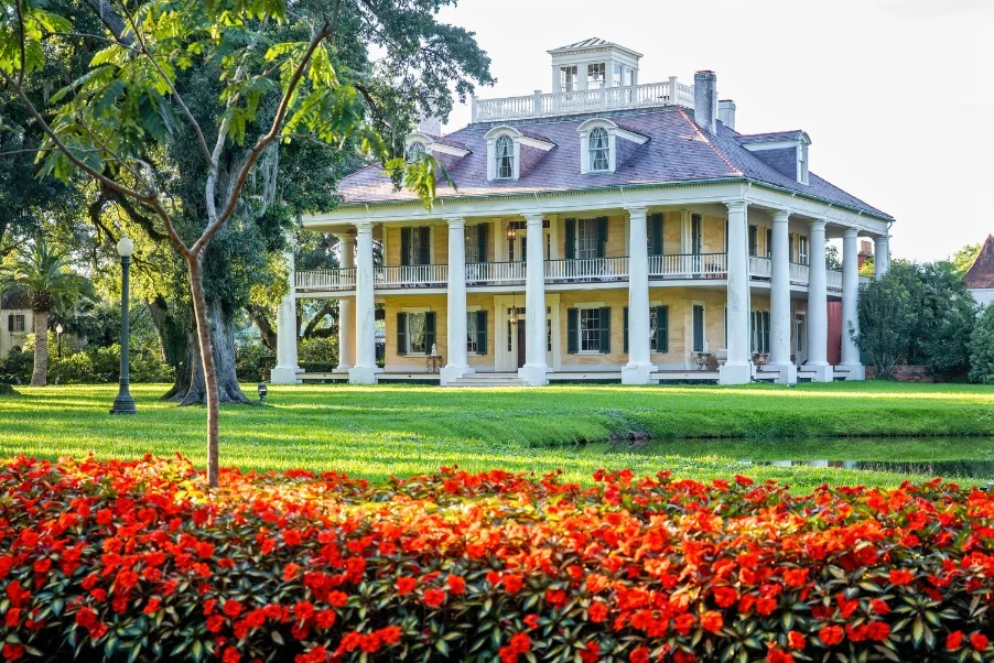 Een klassiek plantagehuis met witte zuilen en dubbele veranda's, omgeven door groen gazon en felrode bloemen bij New Orleans