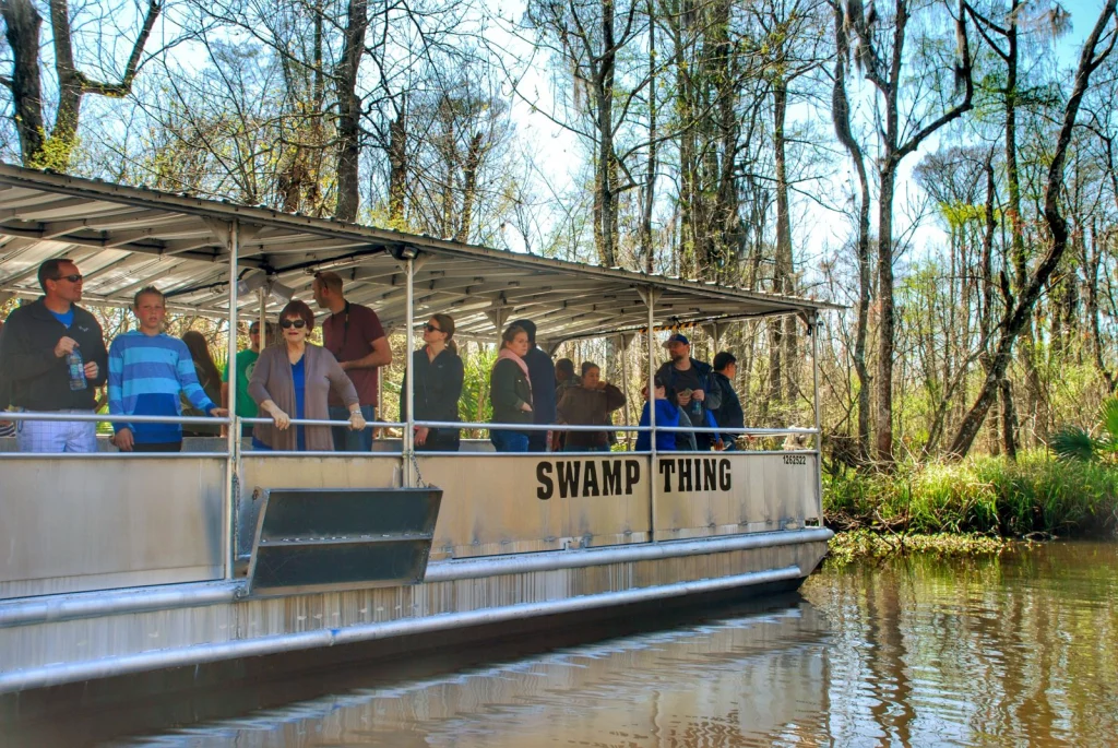 Groep toeristen staat aan de reling van een overdekte rondvaartboot met de naam Swamp Thing tijdens een tocht door een moeras met kale bomen