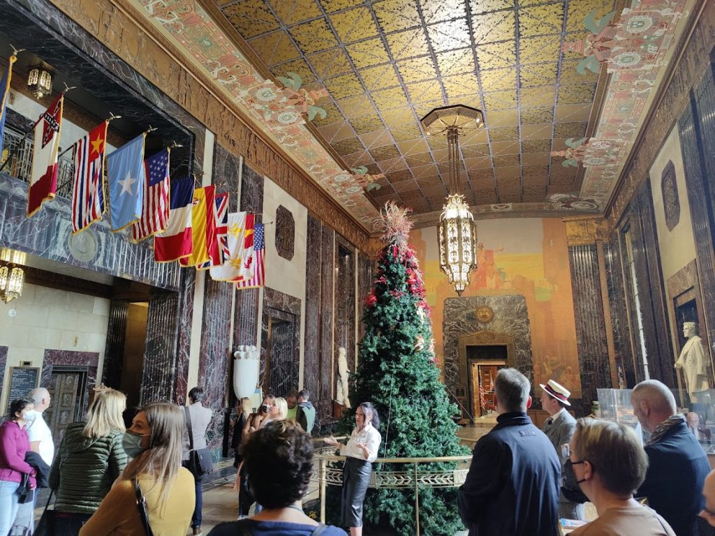 Groep bezoekers bekijkt een sierlijke zaal met goudkleurig plafond, marmeren wanden, hangende vlaggen en een versierde kerstboom